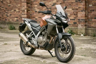Honda CB 500X d'occasion en vue trois quarts avant sur béton urbain, détails d'usure visibles sur le pot d'échappement et les repose-pieds